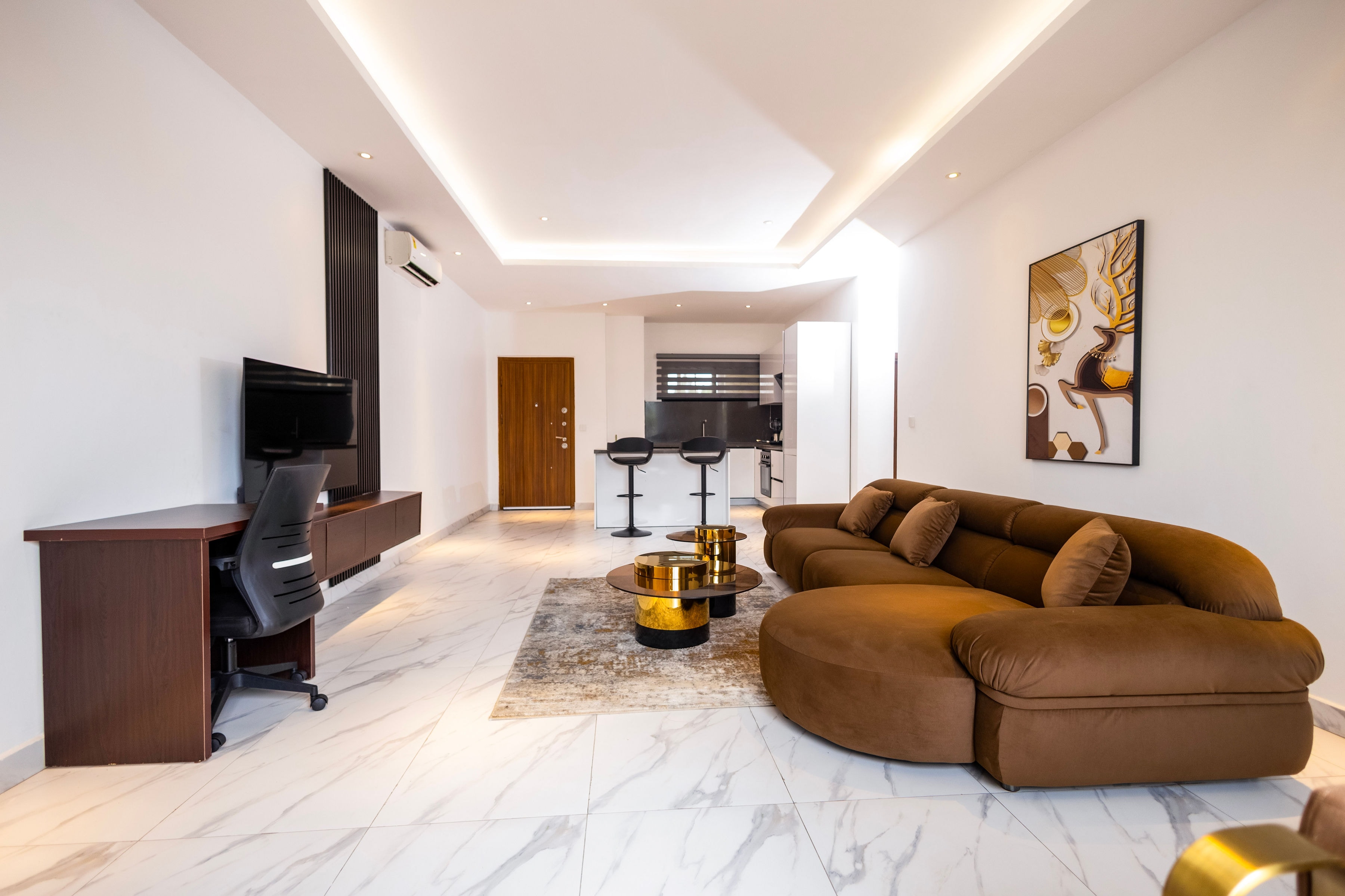 One-bedroom living room with a curved tan sectional, a desk and the kitchen at the far end