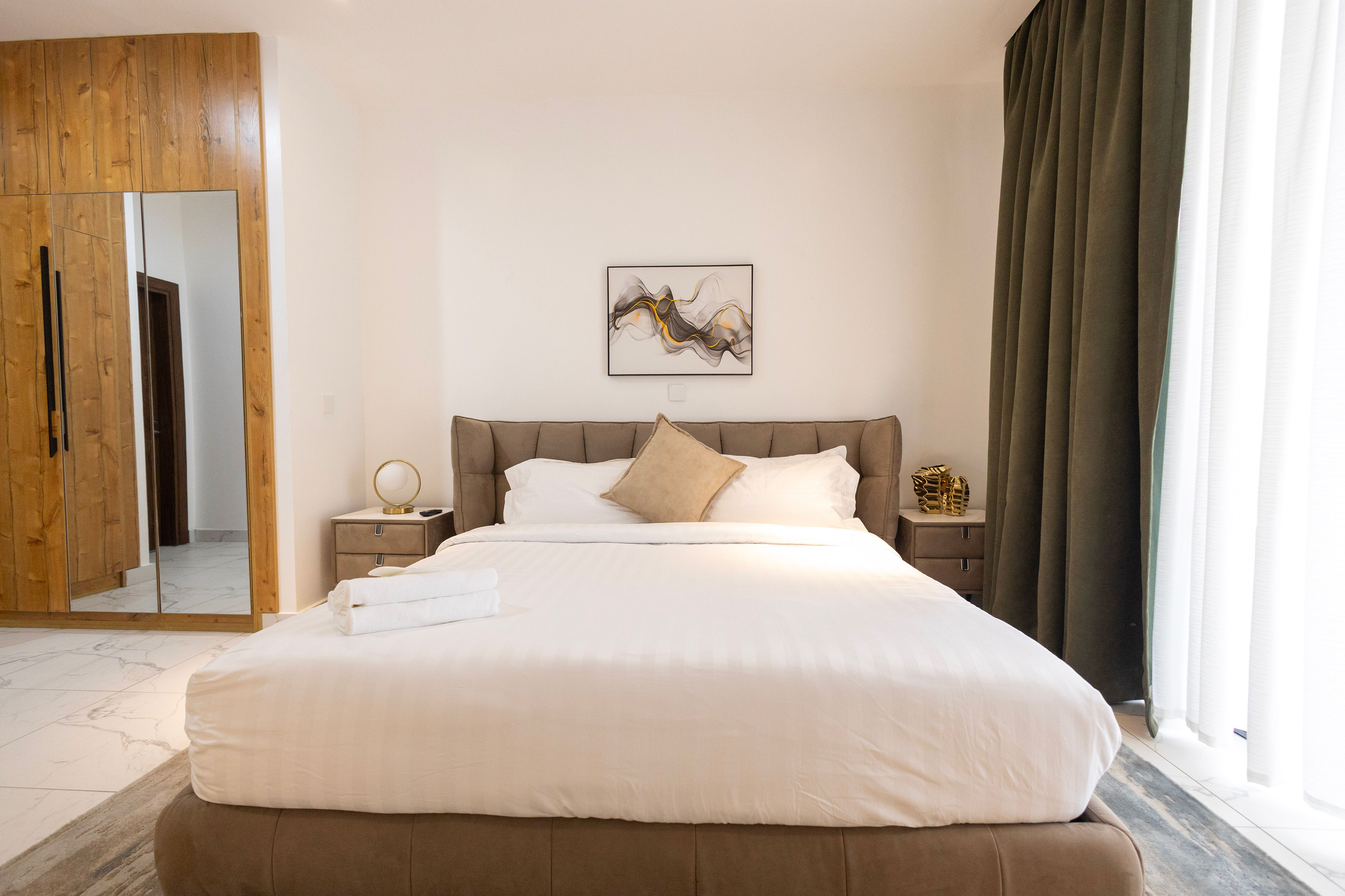 One-bedroom with a tan upholstered bed, timber wardrobe and green curtains